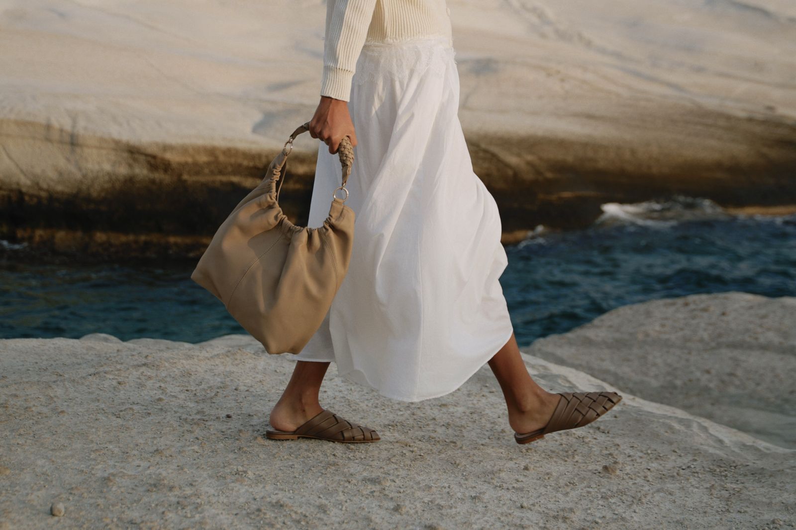 Modelo caminando junto al mar con la Milos Bag Beige y mules tejidas